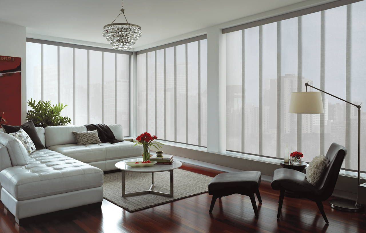 Living room with a sectional sofa, coffee table, and lounge chair surrounded by large windows with panel shades.